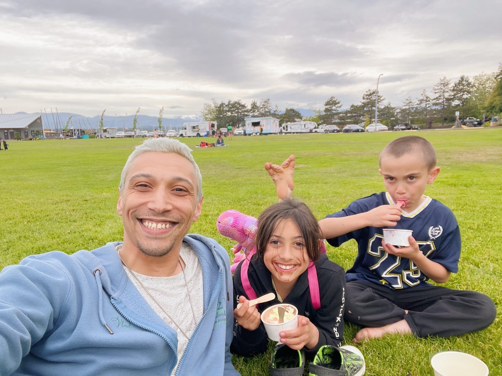 Vlad Pereira and his two kids sitting on the grass at a park, eating gelato from little cups, a food truck and mountains in the background
